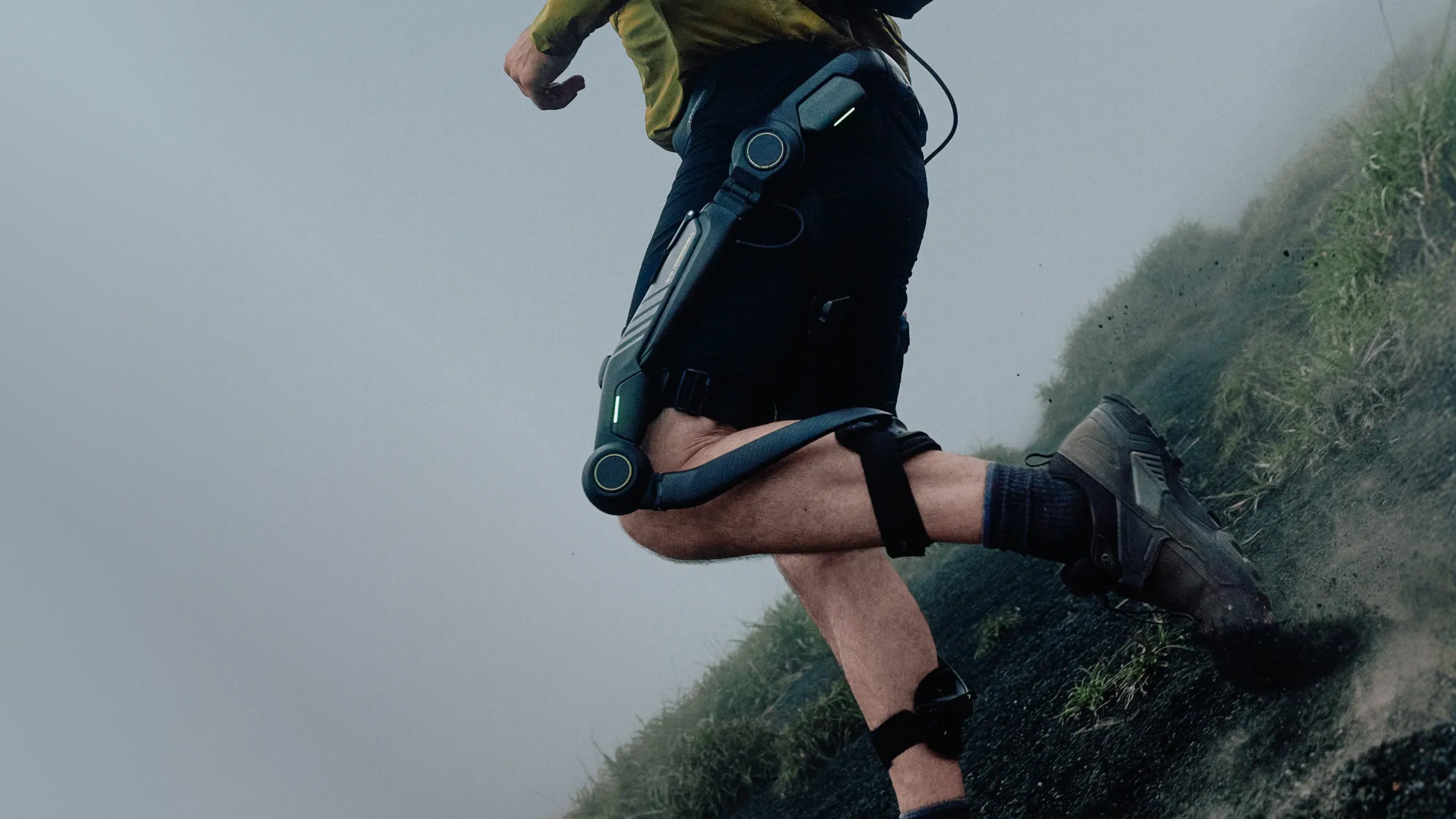 A runner in black athletic shorts and a yellow top wearing a dark blue and black robotic knee exoskeleton while running up a grassy, foggy hillside.