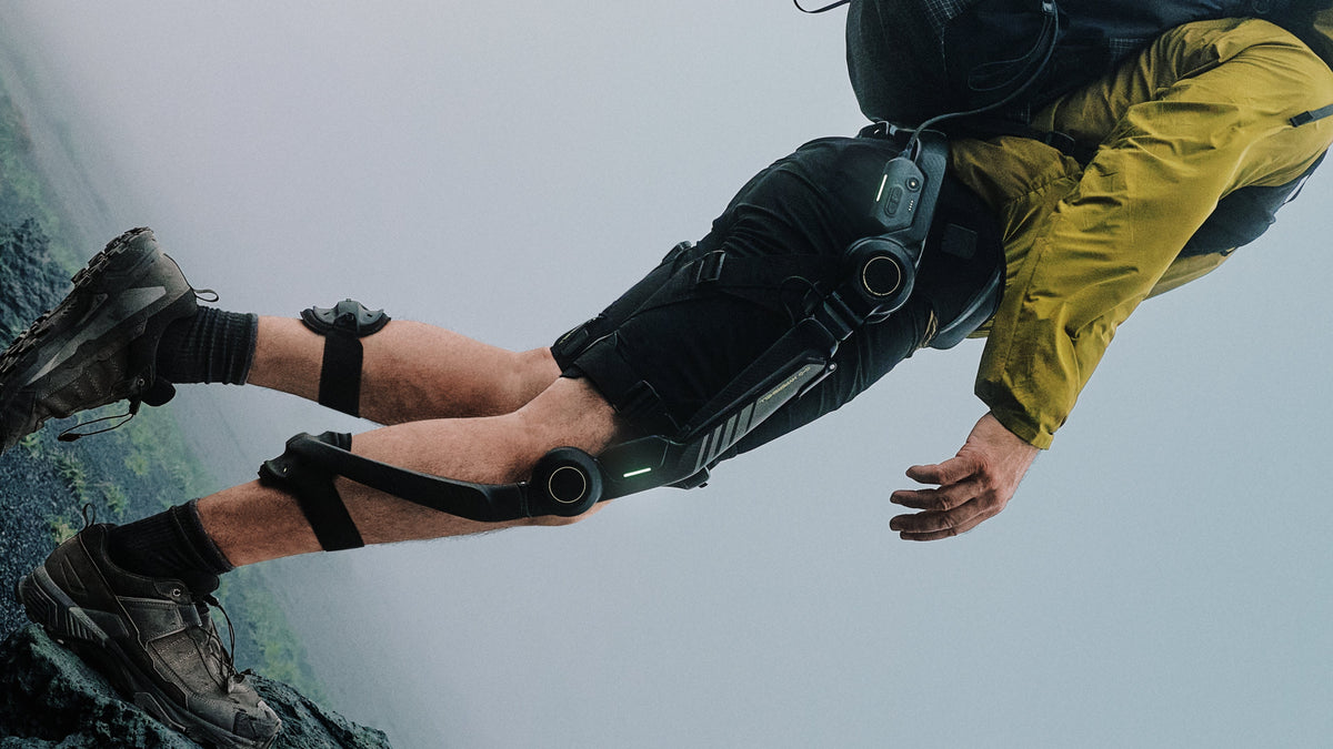 A person outdoors wearing a black robotic exoskeleton leg brace with knee and hip joints, paired with black shorts and a yellow jacket against a misty gray background.